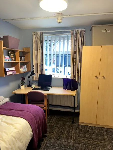 A bedroom in City Campus standard accommodation: a desk with an office chair and desktop computer looking out through a window with a view of car parking, a wardrobe on the right-hand side of the room