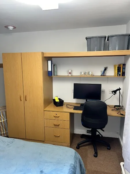 A bedroom in City Campus ensuite accommodation: a desk with attached drawers, wardrobe and set of shelves against the wall to the left of a bed