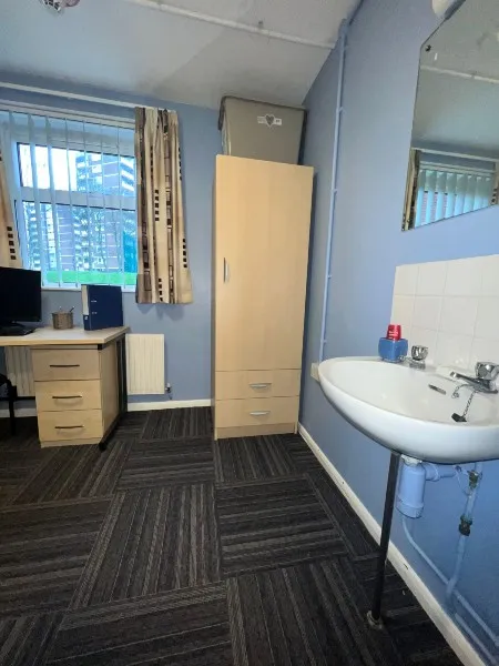 A bedroom in City Campus standard accommodation: a sink unit and mirror on the right side of the room, adjacent to a wardrobe, a desk and chair on the far side of the room looking out through a window towards another accommodation building