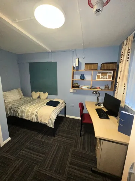 A bedroom in City Campus standard accommodation: a bed adjacent to a computer desk with an office chair, shelves and a pin board on the wall to their left