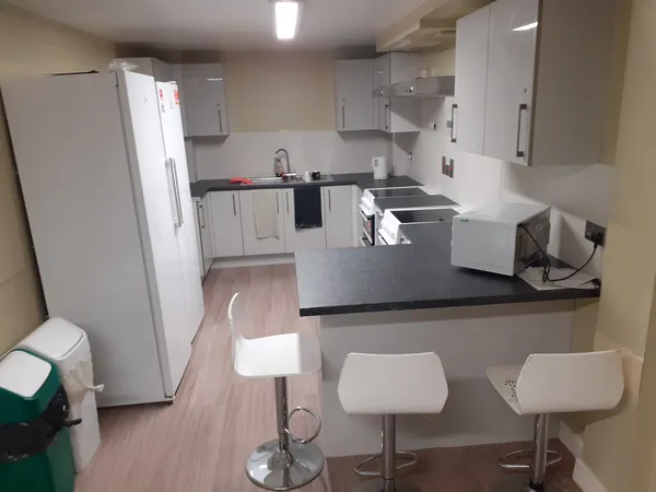 Photograph of a communal kitchen in Walsall Campus accommodation: a seating area, cupboards, ovens, a sink, two dustbins and a fridge-freezer.