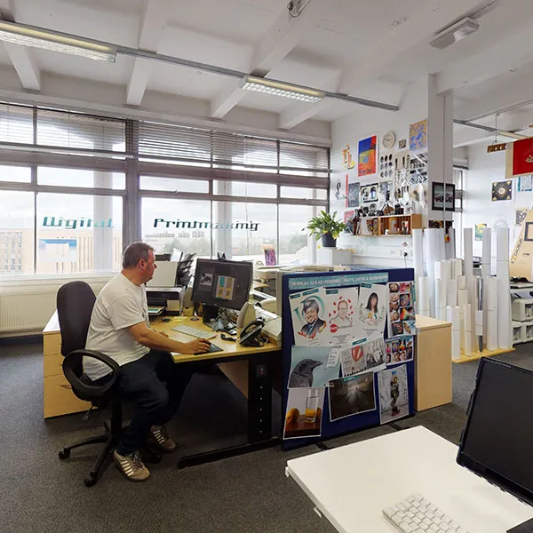 Principal Technician Jim Abernethy working at a desk in the digital Print Support Hub, surrounded by various printed materials, posters, and large-format printers. The workspace is filled with natural light from large windows, and the walls are decorated with colourful artwork and photographs.