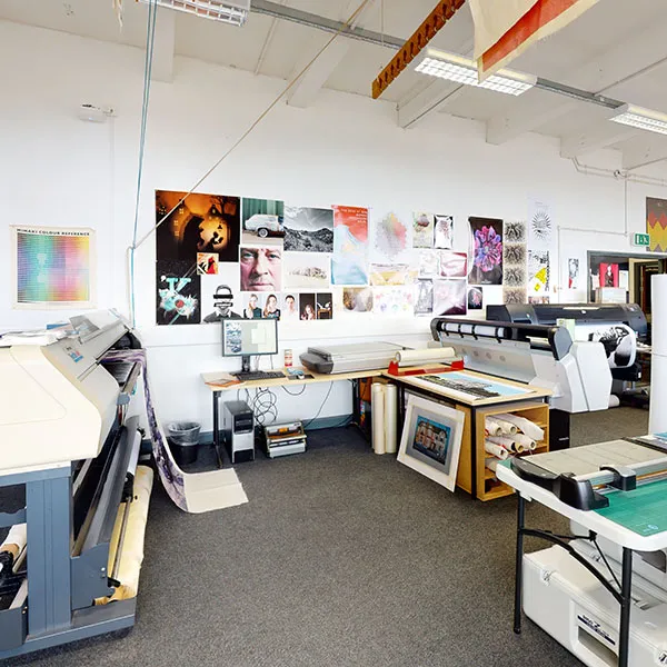 Interior of a digital printmaking studio with large-format printers, worktables, and rolls of paper. The walls are adorned with colorful posters and artwork, showcasing a variety of designs and prints. A cutting mat and tools are set up on a table, ready for use.