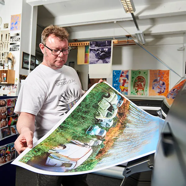 Principal Technician Jim Abernethy holding a large, colourful print of vintage cars in a digital printmaking studio. He is inspecting the print as it comes out of a large-format printer. The studio walls are decorated with various posters and artwork, Jim is wearing a white t-shirt with a graphic design.