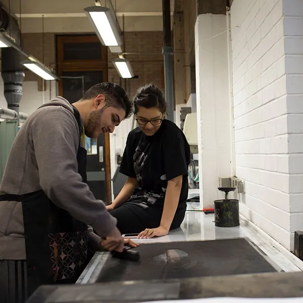 male and female student inking up an etching plate, ready for printing, in the Intaglio print room