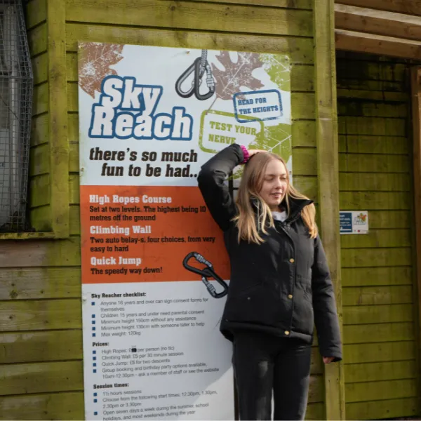 A student standing in front of the Sky Reach sign, advertising the High Ropes Course, Climbing Wall and Quick Jump