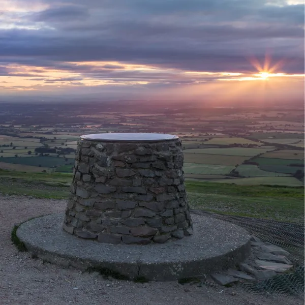 A circular monument made of piled stones in the Wrekin, looking out onto fields and looked upon by a setting sun