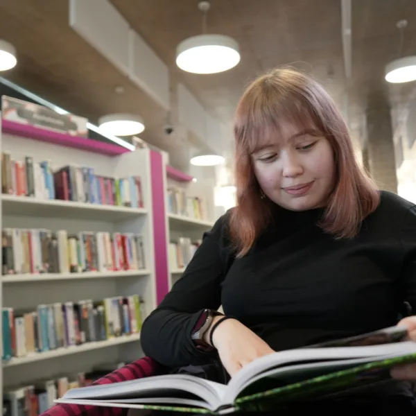 A student in the library sitting in front of bookshelves and reading from a large hardback book