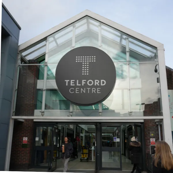 Entrance to Telford Centre shopping centre, a glass wall with a sloped glass roof and a circular sign with a large T logo
