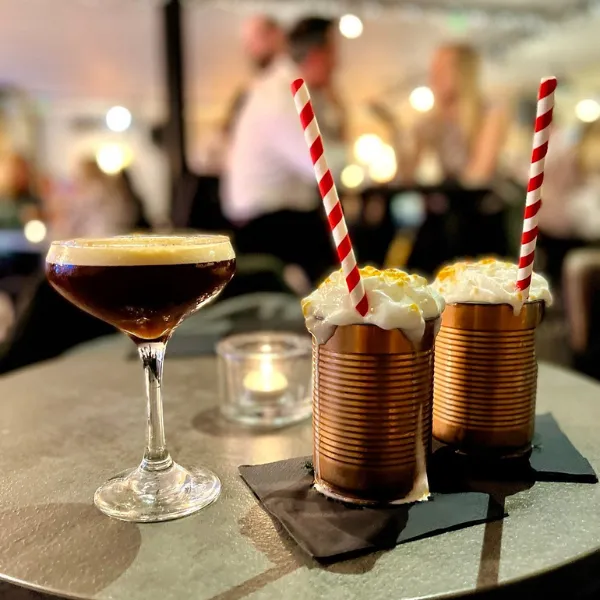 Three drinks, one in a cocktail glass, two in metal cans overflowing with cream and prepared with two paper straws, a candle in a glass dish on the table with them and several other seated tables in the background