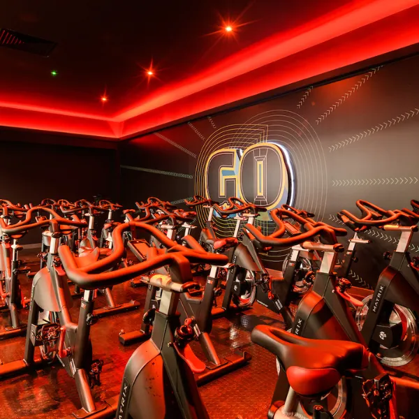 Inside a PureGym, rows of cycling machines illuminated by red lights, a graphic reading GO surrounded by running track lanes on the right wall
