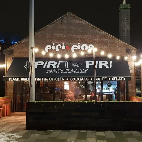 Exterior of Piri Fino restaurant, a fence and square hedge walling in outdoor seating, tarpaulin shelter advertising restaurant specialities: flame grill, piri piri chicken, cocktails, coffee and gelato