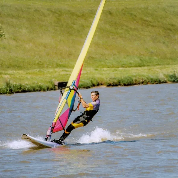 A sailboarder at Sneyd Water Activity Centre, turning their board at a leftwards angle, wearing a yellow and indigo wetsuit