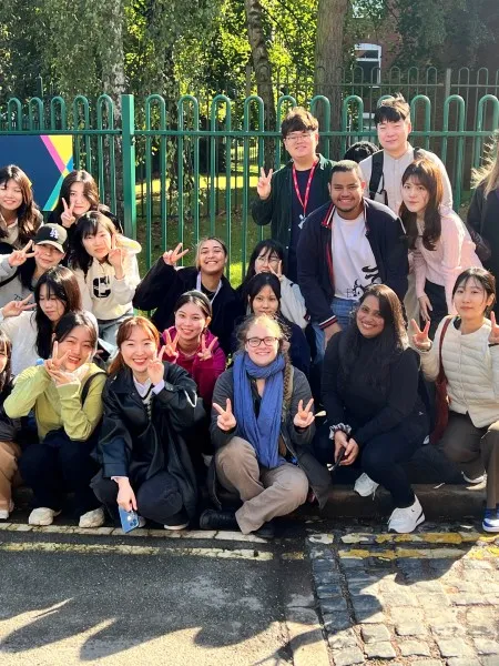 A group of students sitting and standing in a crowd outside the Accommodation Hub reception, facing the camera with smiles and making peace signs