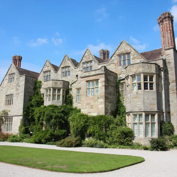Exterior of Benthall Hall: sloping roofs and tall chimney-stacks, foliage growing across the wall of the building, a neat patch of grass partitioning the footpath